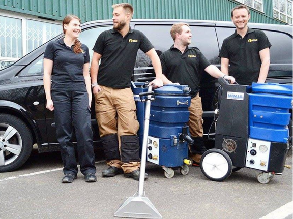 This is the team at K&S Carpet Cleaning, standing together in front of one of their vans, with two carpet cleaning machines in front of them.