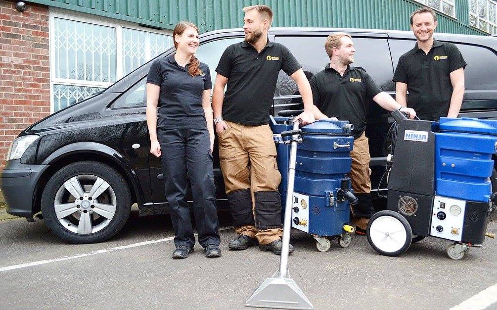 This is the team at K&S Carpet Cleaning, standing together in front of one of their vans, with two carpet cleaning machines in front of them.